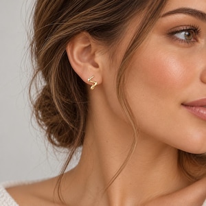 May include: A small gold stud earring featuring a delicate, wavy, serpentine shape. The earring is worn in the earlobe, showcasing a polished metallic finish with a smooth, curved design that follows a subtle, irregular zigzag pattern.