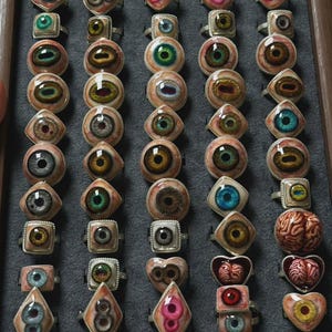 Puede incluir: Una colección de anillos dispuestos en una bandeja de madera, con diversos diseños de ojos con iris realistas en colores como azul, verde, marrón y rosa, junto a dos anillos con forma de cerebros anatómicos, todos montados en marcos de metal.