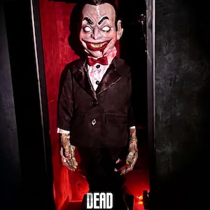 May include: A ventriloquist dummy with pale skin, a wide grin, and white eyes, wearing a black suit, white shirt, and red bow tie. The figure stands inside a dark frame with red lighting. The text DEAD SILENCE and No.57 are visible.