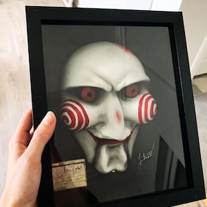 Może przedstawiać: Oprawiona dekoracja ścienna 3D przedstawiająca białą maskę marionetki z czerwonymi spiralnymi policzkami, czerwonymi oczami i czerwonymi ustami. Maska znajduje się na ciemnym tle w czarnej ramie. W lewym dolnym rogu umieszczono kartkę Field Notes.