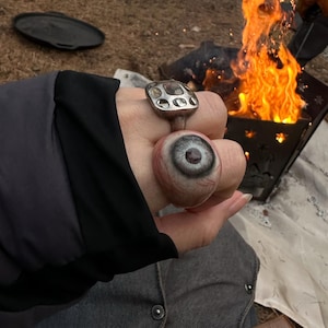 May include: A silver-toned ring featuring a square setting with multiple circular indentations and a second ring designed as a realistic human eyeball with a grey iris, visible veins, and a dark pupil, worn on adjacent fingers.