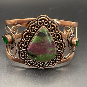 Può includere: Un bracciale a fascia in rame con una grande pietra di zoisite a forma di pera, caratterizzata da tonalità verdi e magenta al centro. La pietra è incorniciata da una montatura in filigrana metallica scura. Due pietre verdi rotonde ai lati.