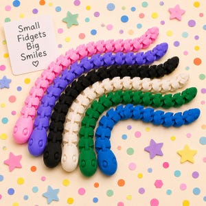 May include: A collection of six articulated snake-shaped fidget toys in pink, purple, black, white, green, and blue. A white rectangular card is included with the text Small Fidgets Big Smiles and a heart symbol printed in black ink.