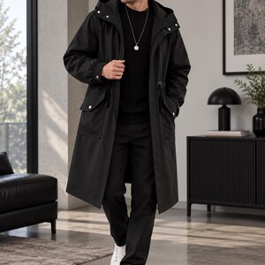 May include: A long black hooded coat featuring a front zipper closure, snap button details, and two large flap pockets at the waist. The coat is worn over a black ribbed sweater and black trousers, paired with white sneakers and a silver pendant necklace.