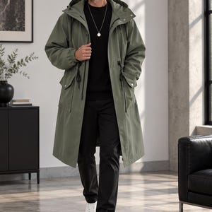 May include: An olive green hooded long coat with a drawstring waist, front flap pockets, and a zipper closure. The coat is worn over a black long-sleeved shirt, black trousers, and white sneakers, with a silver pendant necklace visible at the neck.