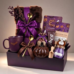 May include: A gift set in a purple box containing a brown faux fur blanket with a purple ribbon, a purple mug with a bat and spiderweb design, two gold spoons, a pumpkin-shaped candle holder, bath salts, tea, a face mask, and shea butter.