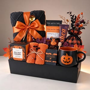 May include: A gift basket containing a dark grey knit blanket with an orange ribbon, a Tea With Tae tea bag, a jar of honey, a tube of shea butter, orange towels, a Sea Salt Caramel cookie box, oatmeal soap, and a black pumpkin mug.