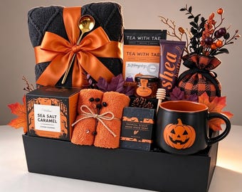 Könnte beinhalten: Ein Geschenkkorb mit einer dunkelgrauen Strickdecke mit orangefarbenem Band, einem Tea With Tae Teebeutel, Honig, Sheabutter, orangefarbenen Handtüchern, Sea Salt Caramel Keksen, Haferflockenseife und einer schwarzen Tasse mit Kürbismotiv.