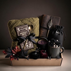 May include: A gift box containing an olive green knit blanket, a chocolate bar labeled Midnight Brew, a gold spoon, a brown leather journal with a skeleton hand pen, a black drawstring pouch, a purple candle, and a dark gray mug with skulls.