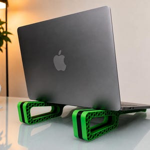 Könnte beinhalten: Ein dunkelgrauer Laptop mit einem Apple-Logo auf dem Deckel steht auf einem leuchtend grünen Laptopständer. Der Ständer weist an den Seiten ein ausgeprägtes Wabenmuster und einen schwarzen Mittelstreifen auf, der den Laptop anhebt.