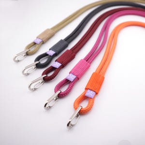 May include: Five braided nylon lanyards in tan, black, dark red, pink, and orange. Each lanyard features a silver metal carabiner clip at the end and a small purple fabric tag with a white logo graphic attached to the cord near the clasp.