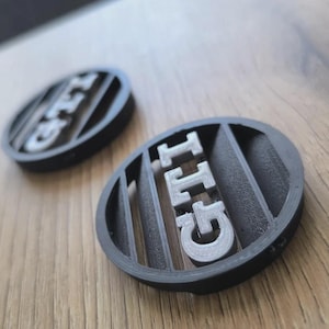 Può includere: Due emblemi circolari in plastica nera con le lettere GTI in rilievo con finitura argentata. Ogni emblema presenta una serie di lamelle orizzontali dietro la scritta, creando un aspetto a griglia. Il design è coerente su entrambi i pezzi.