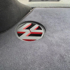 Könnte beinhalten: Ein kreisförmiges Volkswagen-Logo mit schwarzem Rahmen und rotem Hintergrund, eingelassen in eine dunkelgraue Teppichoberfläche. Das Emblem zeigt die ikonischen VW-Buchstaben im Ausschnitt-Stil und befindet sich auf einer Hutablage.