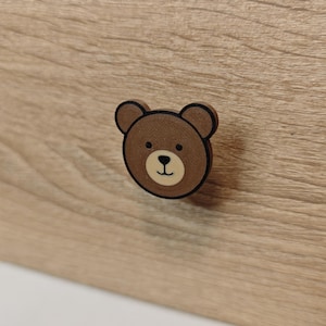 May include: A brown bear-shaped drawer knob with a light beige muzzle, black eyes, and a black nose. The bear face has a dark brown outline and is mounted on a light wood-grain drawer front. The knob features a flat, circular design.