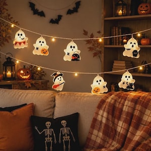 Peut inclure: Une guirlande lumineuse LED pour Halloween composée de caches en forme de fantômes blancs. Chaque fantôme présente un motif unique, incluant un seau citrouille, un chat noir, une pierre tombale et un chapeau de sorcière sur un fil blanc.