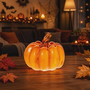 Peut inclure: Une figurine de citrouille en verre orange translucide avec un corps lisse et arrondi et une tige incurvée. La citrouille présente un aspect vibrant et lumineux, posée sur une surface en bois, entourée de feuilles d'automne rouges et dorées.
