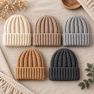May include: Five knit beanies arranged in two rows, featuring a ribbed texture and a folded cuff. The colors include cream, light tan, light gray, medium brown, and dark charcoal gray. Each hat has a consistent vertical stitch pattern throughout.