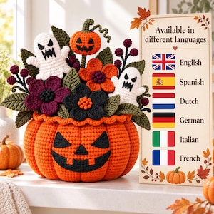 Może przedstawiać: Szydełkowa dekoracja na Halloween z dużą pomarańczową dynią z czarną twarzą jack-o'-lantern, wypełniona dwoma białymi duchami, małą dynią oraz ciemnofioletowymi i pomarańczowymi kwiatami z zielonymi liśćmi.
