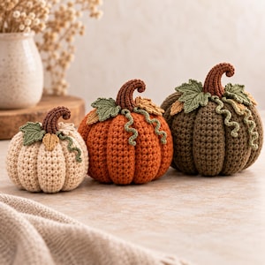 Puede incluir: Tres calabazas tejidas a ganchillo de diferentes tamaños y colores, incluyendo crema, naranja y verde oliva. Cada calabaza presenta un tallo marrón oscuro, hojas verdes tejidas y pequeños zarcillos rizados sobre los cuerpos texturizados.