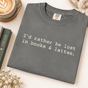 Peut inclure: Un t-shirt gris anthracite à col rond avec le texte imprimé en blanc "I'd rather be lost in books & lattes.". Le t-shirt présente une étiquette de marque Comfort Colors à l'intérieur du col, indiquant une taille large, en coton uni.