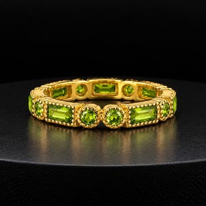 Könnte beinhalten: Ein goldener Bandring mit einem abwechselnden Muster aus rund und im Baguetteschliff geschliffenen grünen Peridot-Edelsteinen. Die Metallfassung hat eine perlenartige Textur um jeden Stein, die ein durchgehendes Design bildet.