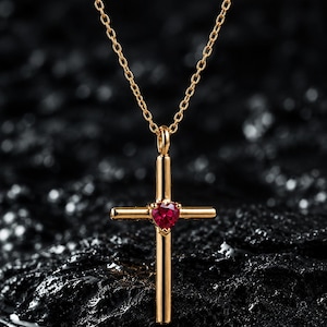 Könnte beinhalten: Eine goldfarbene Kreuz-Halskette mit einem kleinen, facettierten, herzförmigen roten Edelstein, der in der Mitte des Kreuzes eingesetzt ist. Der Anhänger hängt an einer zarten, goldfarbenen Kette mit polierter, reflektierender Oberfläche.