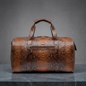 Könnte beinhalten: Eine braune Reisetasche mit durchgehendem, strukturiertem Schlangenhautmuster. Die Tasche verfügt über zwei Tragegriffe, eine horizontale Reißverschlusstasche auf der Vorderseite und einen Hauptreißverschluss an der Oberkante in zylindrischer Form.