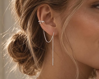 May include: A silver-toned ear cuff featuring a delicate, thin chain that drapes from the cuff to a straight, slender bar earring. The ear cuff has a small, crisscross design, and the entire piece is crafted from a polished, metallic material.