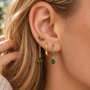 May include: Three gold-toned earrings worn in an earlobe. One earring features a green crystal leaf stud with a dangling chain and teardrop pendant. A plain gold hoop sits in the middle, and a green crystal-encrusted hoop with a teardrop charm.