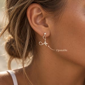 May include: A silver-toned earring shaped like a pair of scissors with a small ball stud at the top. The earring features a delicate design with curved handles and pointed blades. The text "Openable" is printed next to the earring.