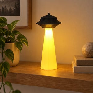 Puede incluir: Una lámpara de mesa con forma de platillo volante con una sección superior negra y una base cónica que emite una luz amarilla cálida. La lámpara presenta una parte superior puntiaguda y un borde ancho, con el haz de luz extendiéndose.