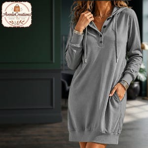 Puede incluir: Un vestido tipo sudadera con capucha de color gris que presenta mangas largas con puños acanalados, una tapeta de botones parcial en el escote, una capucha con cordón y dos bolsillos laterales. La prenda tiene un dobladillo recto y un color gris sólido.
