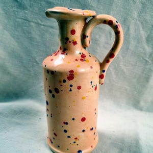 Puede incluir: Una jarra de cerámica con un esmalte de color melocotón claro que presenta un patrón moteado de salpicaduras de pintura roja, amarilla y azul. El recipiente tiene un cuerpo cilíndrico, un cuello estrecho con borde y un asa curva lateral.