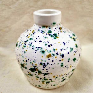 Könnte beinhalten: Eine kleine, runde Keramikvase mit weißer, glänzender Oberfläche, die mit einem gesprenkelten Muster aus dunkelblauen, waldgrünen und senfgelben Farbspritzern überzogen ist, mit einem kurzen, schmalen Hals und einem bauchigen Körper.