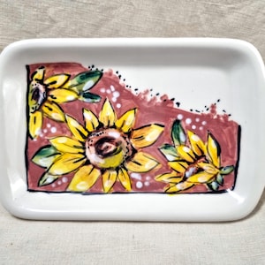 Puede incluir: Una bandeja de servicio rectangular de cerámica blanca con asas laterales integradas. El interior presenta un diseño pintado a mano de tres girasoles amarillos sobre un fondo malva apagado, acentuado con puntos blancos y contornos negros.