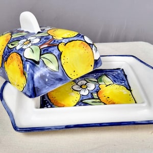 Può includere: Un burriera in ceramica con coperchio rimovibile, caratterizzata da un motivo dipinto a mano di limoni giallo brillante, foglie verdi e fiori bianchi su uno sfondo blu intenso. La base rettangolare presenta un bordo bianco e una finitura blu.