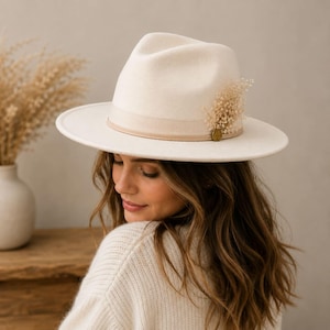 May include: A cream-coloured fedora hat featuring a wide brim and a matching tonal ribbon band. The hat is accented with a small cluster of dried beige flowers tucked into the side of the band, secured by a small, circular metallic gold-toned charm.