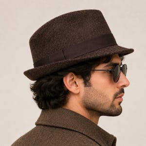 Könnte beinhalten: Ein dunkelbrauner Filz-Fedora-Hut mit einem passenden dunkelbraunen Ripsband. Der Hut verfügt über eine strukturierte Krone und eine leicht nach unten geneigte Krempe, was für eine klassische Silhouette sorgt. Die Filzstruktur ist sichtbar.