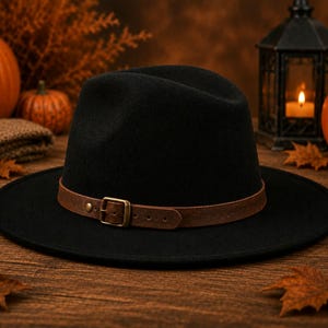 Może przedstawiać: Czarny kapelusz typu fedora z szerokim rondem i brązowym paskiem z imitacji skóry wokół główki. Pasek jest zabezpieczony metalową klamrą i małym nitem, co stanowi kontrast dla jednolitego czarnego materiału, z którego wykonano konstrukcję kapelusza.