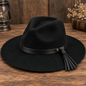 Puede incluir: Un sombrero fedora negro con ala ancha y copa pellizcada. El sombrero presenta una banda de aspecto de cuero negro que rodea la base, acentuada con un remache metálico y un detalle de borla de aspecto de cuero negro colgando en el lateral.