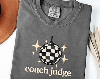 May include: A charcoal gray crew neck t-shirt featuring a graphic of a black and white checkered disco ball surrounded by light-colored star shapes. Below the disco ball, the text reads couch judge in a light-colored, rounded font.