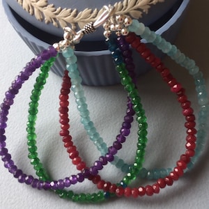 Op de afbeelding: Vier kralenarmbanden in verschillende kleuren: paars, groen, rood en blauw. De armbanden zijn gemaakt van kleine, gefacetteerde kralen en hebben zilveren sluitingen.