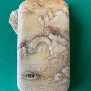 Puede incluir: Un colgante de marfil tallado con una escena detallada de un puente y montañas. El colgante tiene un agujero en la parte superior para colgar.