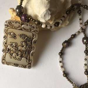 May include: A white jade pendant necklace with a carved design of animals and clouds. The necklace is made with white and purple beads and bronze chain.