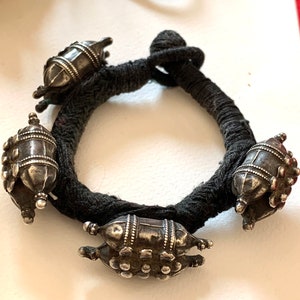 May include: A silver bracelet with four decorative charms, each with a unique design. The bracelet is made of black thread and is tied together with a knot.