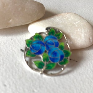 May include: A silver and enamel pin featuring three blue flowers with green leaves. The pin is circular in shape and has a delicate design.