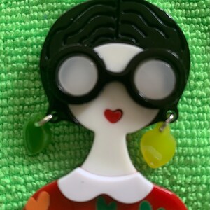 Puede incluir: Una broche de plástico roja, blanca y negra de una mujer con un moño, que lleva gafas y pendientes. La mujer lleva un vestido rojo con un cuello blanco y un patrón de corazones verdes y naranjas.