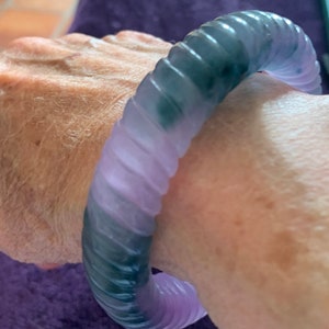 May include: A jade bangle bracelet with a swirling pattern of green and purple. The bracelet is made of a smooth, polished jade and has a slightly rounded shape.