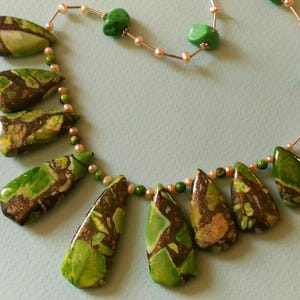 Puede incluir: Un collar de piedras preciosas verdes y marrones con una cadena de plata y pequeñas cuentas blancas. El collar presenta una variedad de colgantes de piedras preciosas verdes y marrones.