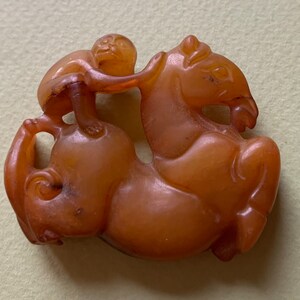 Peut inclure: Un pendentif en ambre sculpté représentant un singe monté sur un cheval. Le pendentif est de couleur orange foncé et a une finition lisse et polie.